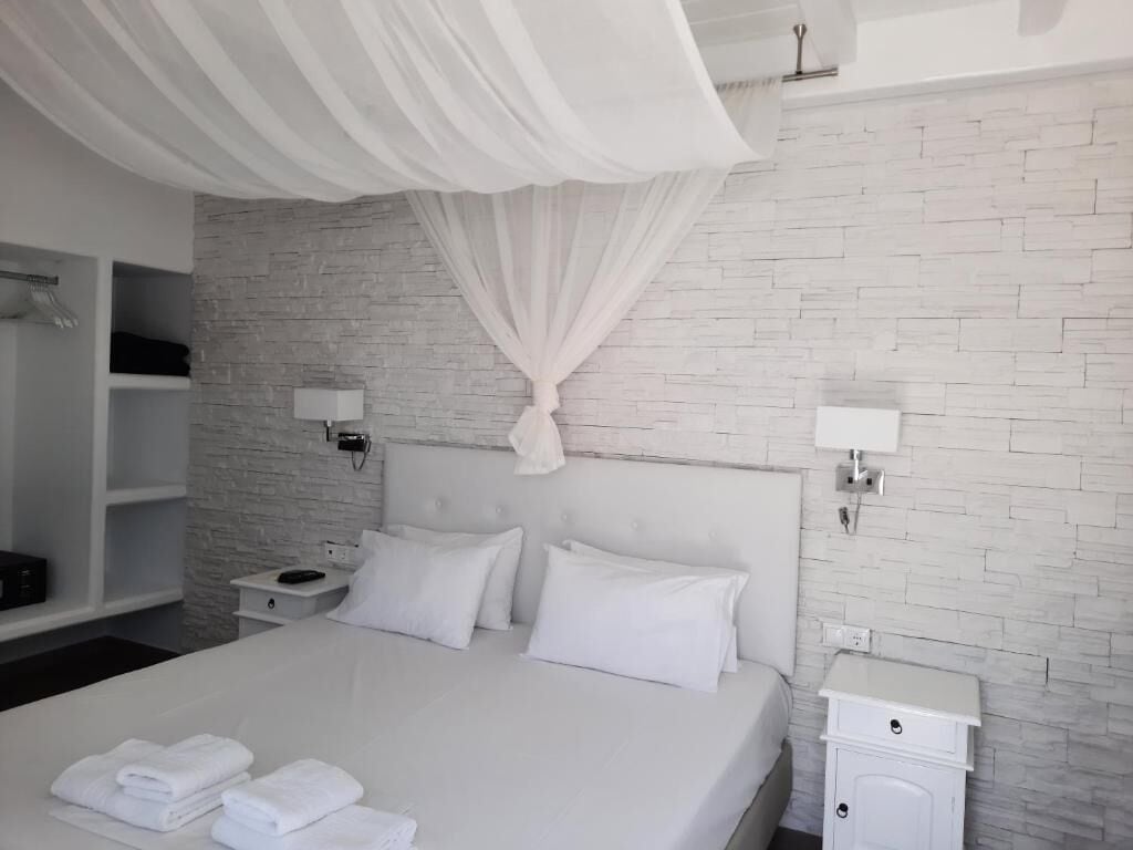 A modern bedroom with a white bed, white curtains, a brick-patterned wall, and a white nightstand with a lamp.