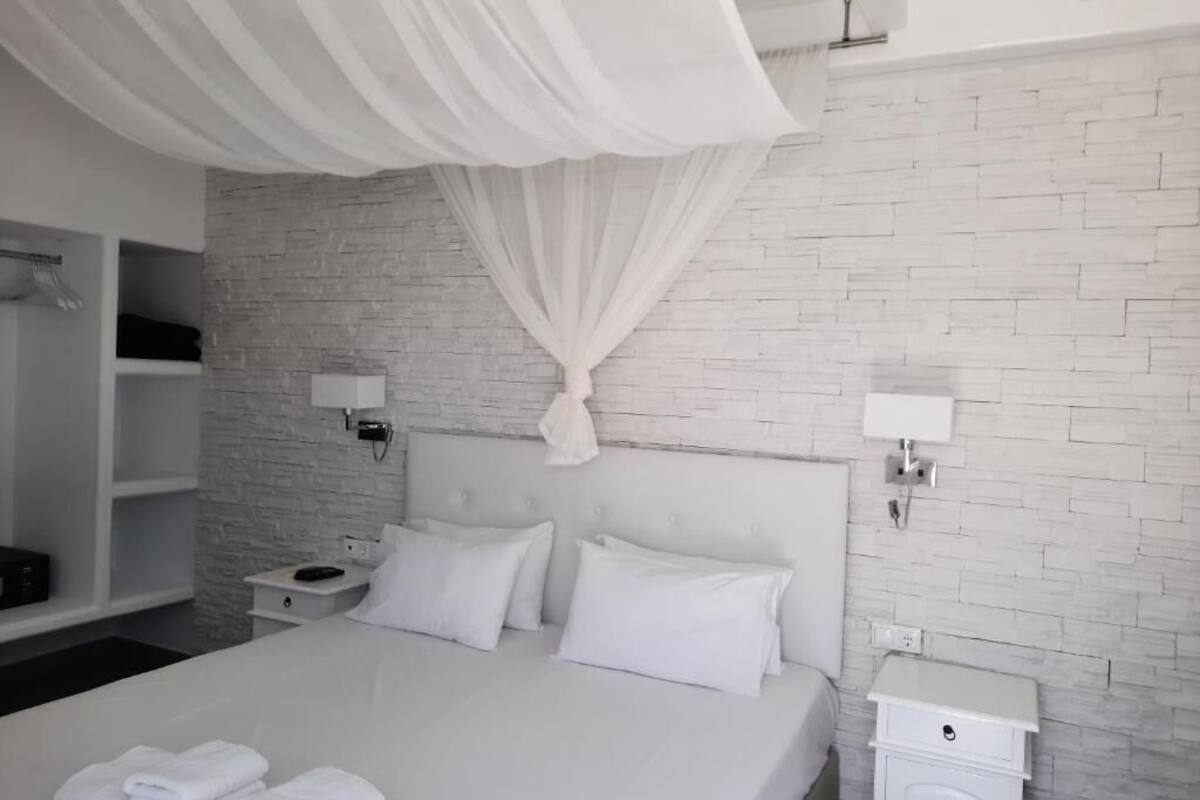 A modern bedroom with a white bed, white curtains, a brick-patterned wall, and a white nightstand with a lamp.