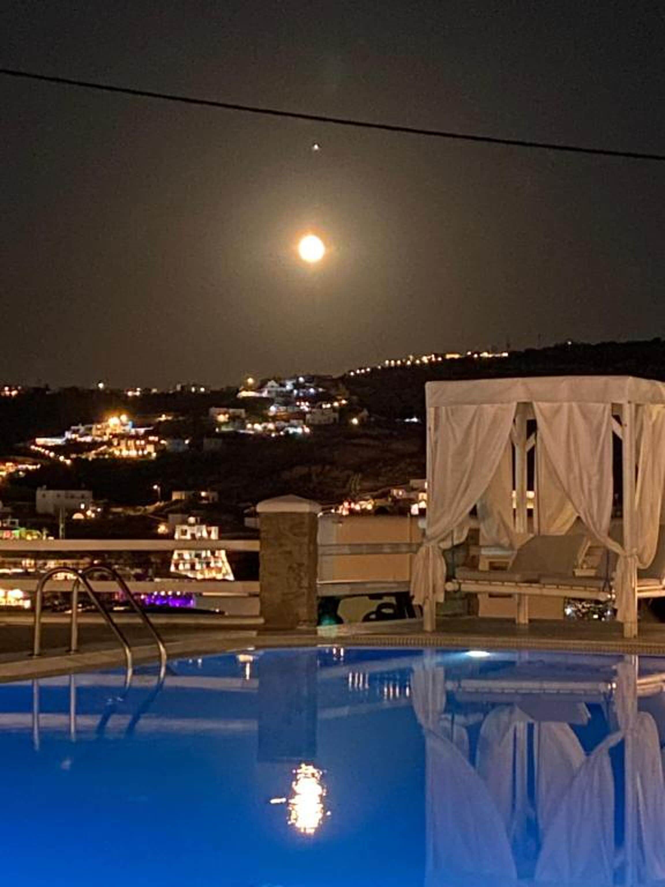 Ein Pool mit Sonnensegel, Blick auf eine Stadt bei Nacht und ein voller Mond am Himmel.
