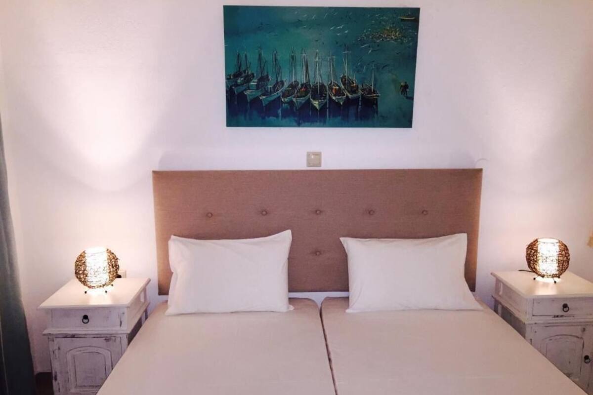 A double bed with white linens, a wooden headboard, two bedside tables with lamps, and a painting of boats on the wall.