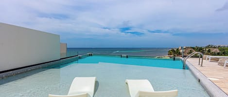 A clear swimming pool with two lounge chairs, a view of the ocean, and a tiled deck area.