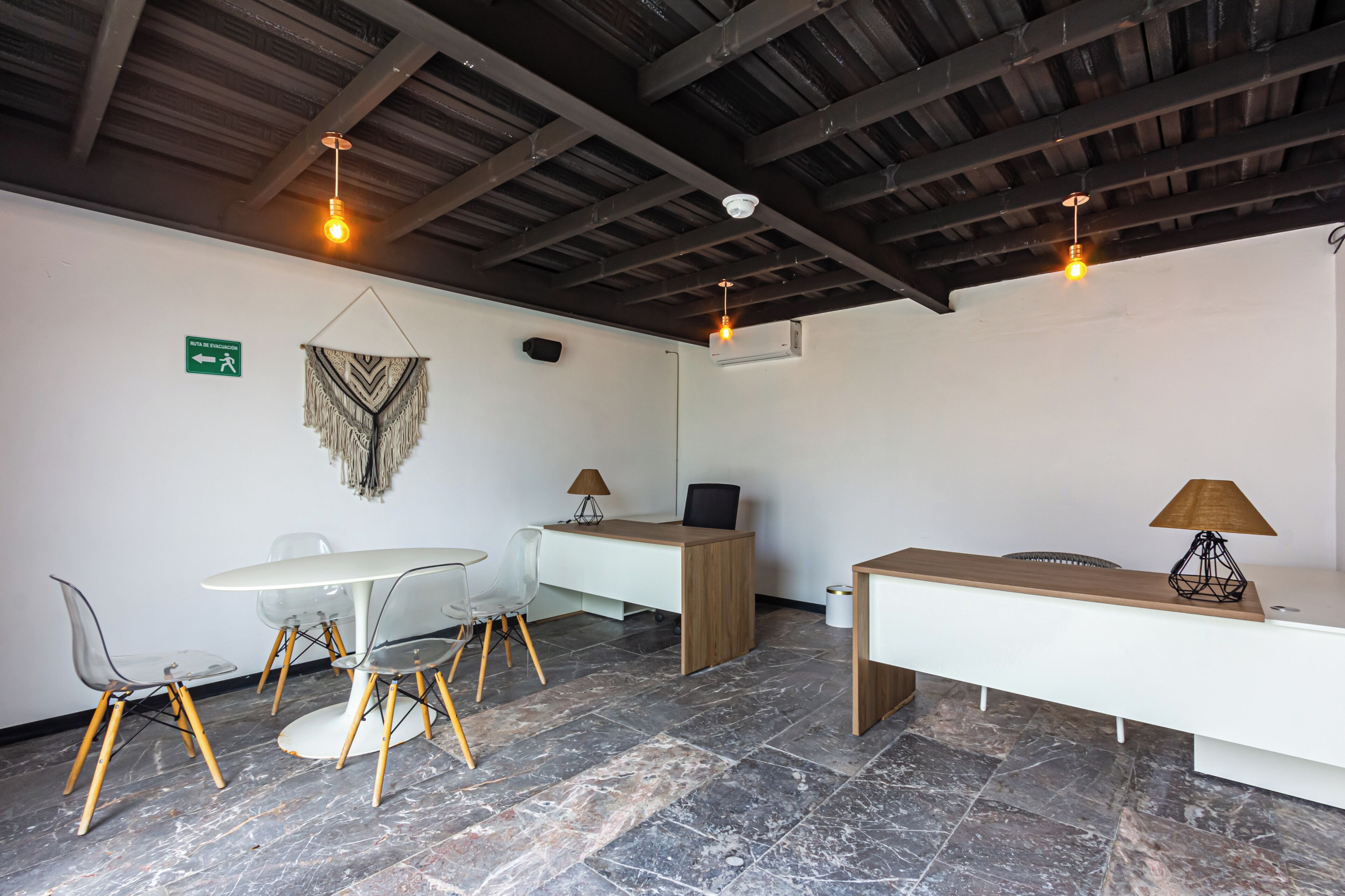 Een moderne kamer met een wit bureau, twee stoelen, een ronde tafel en een bankje. Er zijn plafondlampen en een bord op de muur.