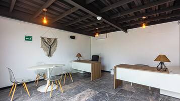 Een moderne kamer met een wit bureau, twee stoelen, een ronde tafel en een bankje. Er zijn plafondlampen en een bord op de muur.