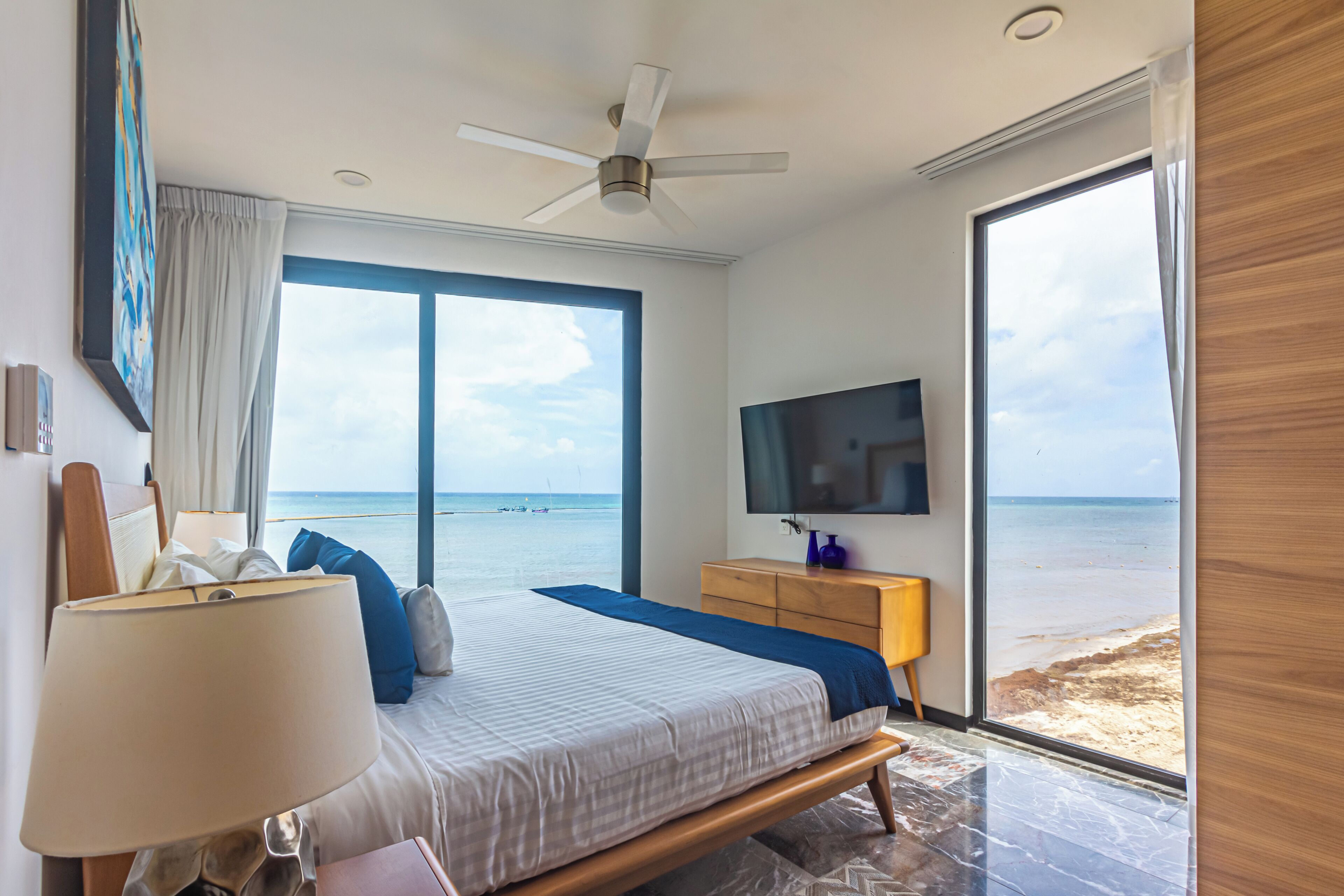 Slaapkamer met een bed, een televisie, een plafondventilator, een raam met uitzicht op het strand en een houten nachtkastje.