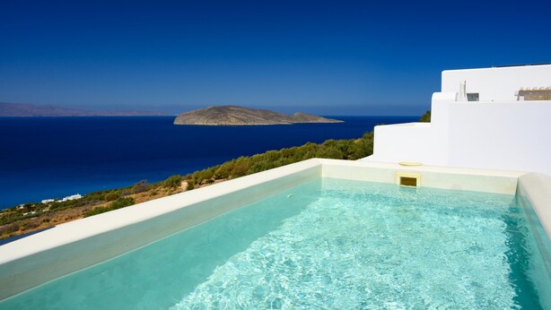 Ein Pool mit klarem, blauem Wasser, ein weißes Gebäude und Blick auf das Meer sowie eine entfernte Insel.