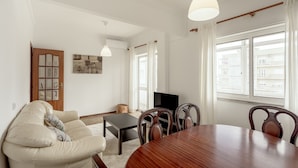 Una sala de estar con sofá, mesa de comedor, sillas, televisor y ventanas amplias.