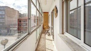 Un pasillo estrecho con ventanas amplias, una puerta de madera y un pequeño rincón de descanso con una mesa y sillas.