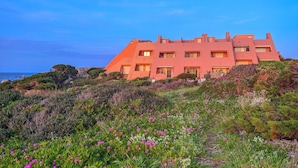 A large, multi-story building with a flat roof, surrounded by a variety of vegetation and flowers. - Casa Audrey by Wonderful Italy (Santa Teresa di Gallura)