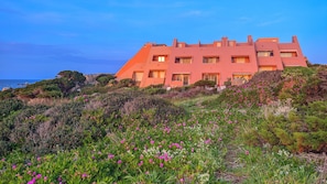 A large, multi-story building with a flat roof, surrounded by a variety of vegetation and flowers. - Casa Audrey by Wonderful Italy (Santa Teresa di Gallura)