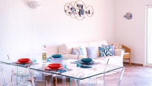 A dining area with a glass table and chairs, a white sofa, and a wall art piece. - Casa Audrey by Wonderful Italy (Santa Teresa di Gallura)