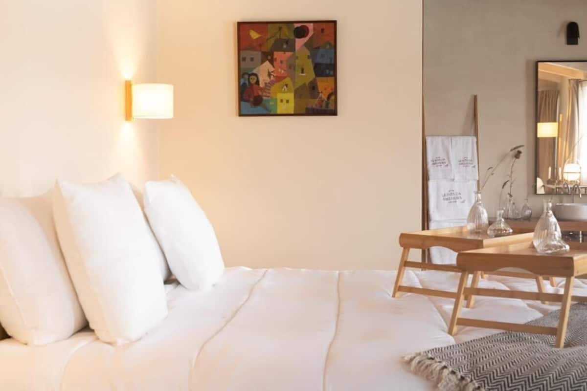 Ett hotellrum med en säng, kuddar, ett sängbord med glas, en tavla på väggen och en spegel.