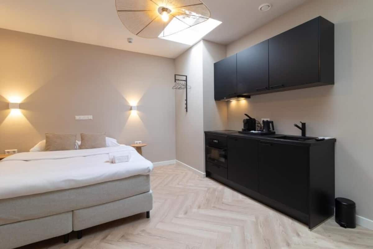 A compact bedroom with a bed, bedside lamps, and a small bedside table. Adjacent is a kitchenette with a stove, sink, and cabinets.