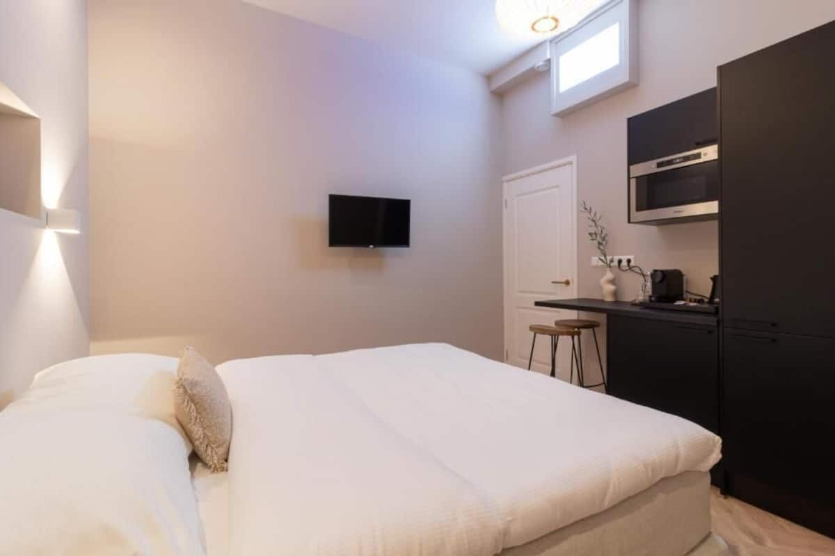 A bedroom with a bed, a TV mounted on the wall, a desk with a chair, and a kitchen area with a microwave and oven.