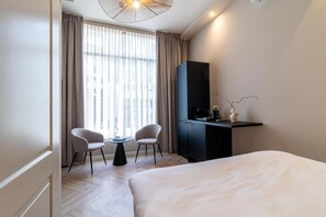 Ein Hotelzimmer mit einem Bett, zwei Sesseln, einem Schreibtisch mit Fernseher und einem Fenster mit Vorhängen.