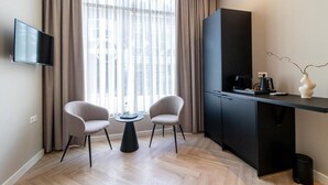 A modern hotel room with a flat-screen TV, two chairs, a small round table, a black cabinet, and a large window with curtains. - SAYŌ Boutique Hotel (The Hague)