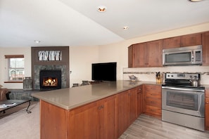 A modern kitchen with a stone fireplace, wooden cabinets, a flat-screen TV, and stainless steel appliances. - Pool|2 Hot Tubs|Garage|Fireplace|Elevator|Balcony (Breckenridge)
