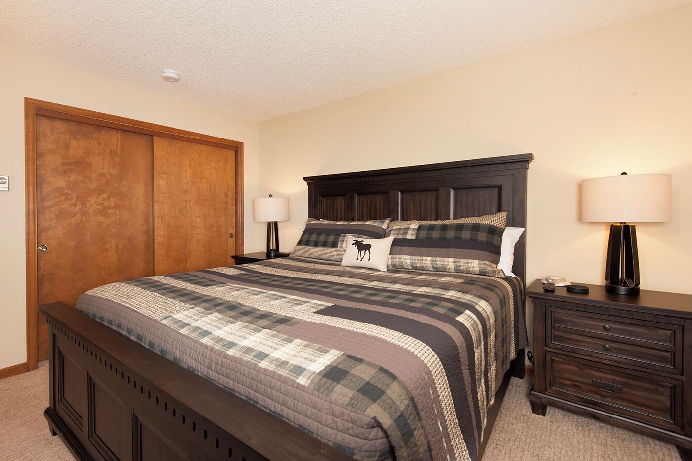 A bedroom with a large bed, a wooden wardrobe, a nightstand with a lamp, and a dresser.