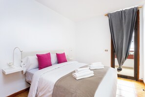 A hotel room with a bed, pillows, a bedside table, a lamp, a window with curtains, and a view of the outside. - Home2Book Cozy Arona Apt with Pool & Terrace (Playa de la Américas)