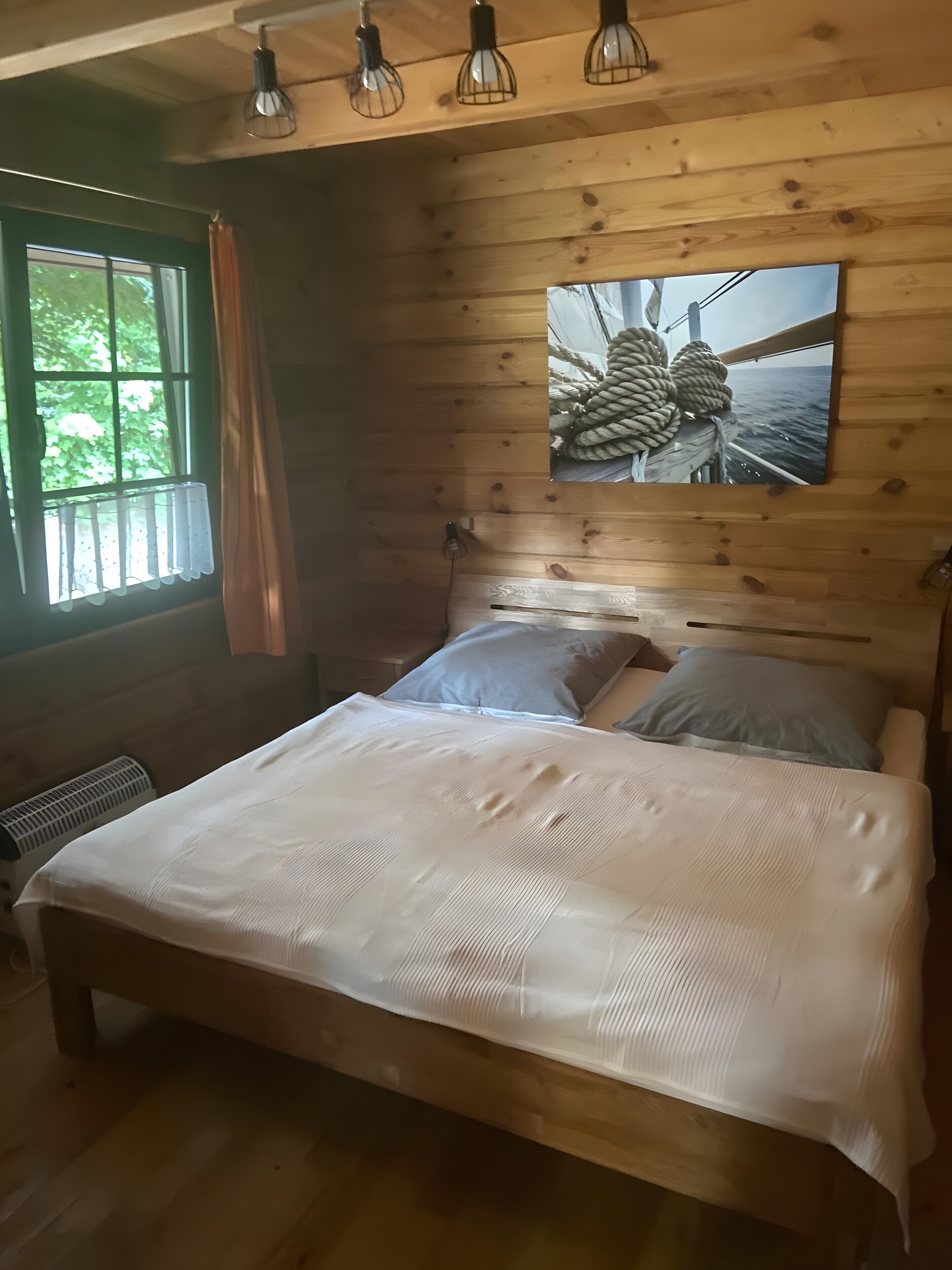 Una habitación tipo cabaña de madera con una cama, una ventana con cortinas, un radiador y un cuadro colgado en la pared.