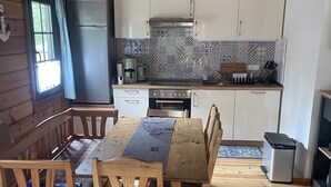 Interior de una cabaña de madera con una mesa de comedor, sillas, una cocina con armarios blancos y un refrigerador.