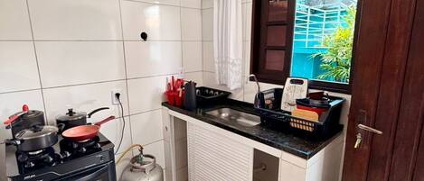 Cocina compacta con estufa a gas, horno y cilindro de gas, un mostrador pequeño con pileta y una ventana con vista a la vegetación.