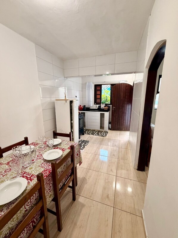 Un comedor con una mesa preparada para cuatro personas, una cocina al fondo y un suelo de baldosas.