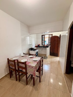 A dining area with a table set for four, a kitchen in the background, and a wooden door. - Pousada Villa Caiçara (Ubatuba)