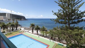Ein Poolbereich mit Blick auf das Meer, Palmen und einem Gebäude im Hintergrund.