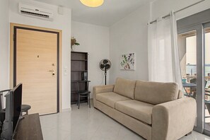 A living room with a beige sofa, a standing fan, a bookshelf, and a door. - Apartment 'Happiness in Chania' with Wi-Fi (Chania)