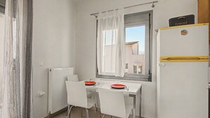 Ett litet, rent rum med ett bord dukat för två personer, ett fönster med gardiner, ett element och ett kylskåp med en skylt som visar att det finns bröd.