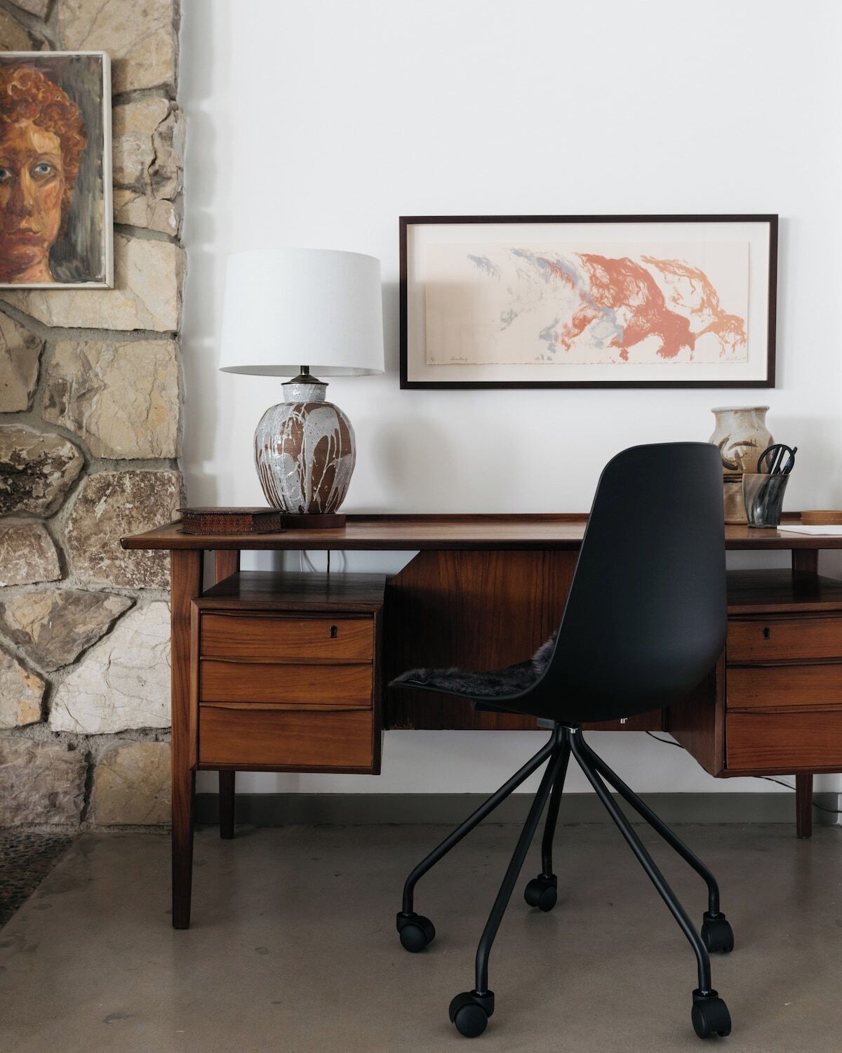 A wooden desk with a black office chair, a stone wall, and a framed abstract painting on the wall.