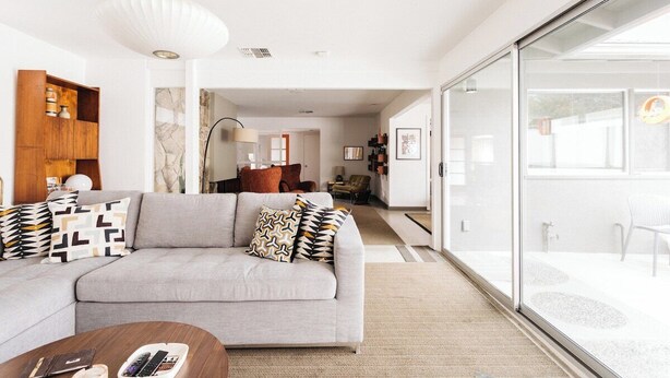 Ein modernes Wohnzimmer mit einem grauen Sofa, gemusterten Kissen, einer Holzkommode und einer großen Glasschiebetür, die zu einem Außenbereich führt.