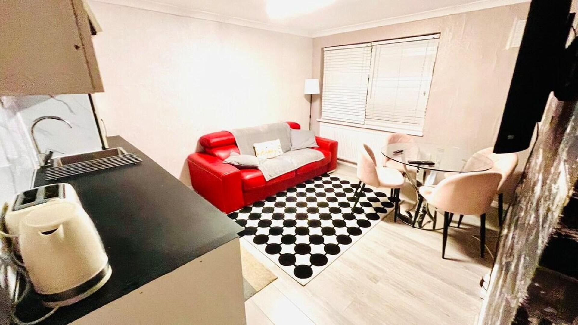 A compact living space with a kitchenette, a red sofa, a small dining table with chairs, and a checkered rug.