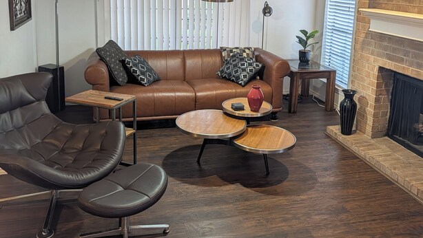 A living room with a brown leather sofa, two round coffee tables, a black leather armchair, a fireplace, and a large window with blinds.