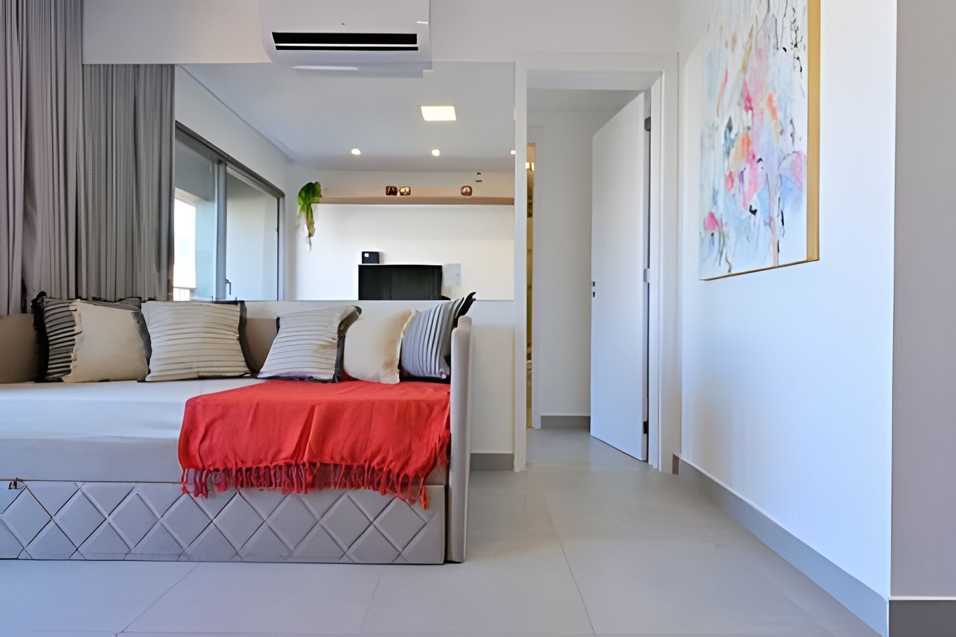 A modern hotel room with a bed, a TV, a painting, and a hallway leading to another room.