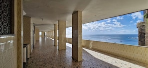 A corridor with patterned walls and a tiled floor, leading to a large window overlooking a body of water with boats. - CODDJ0100 - Cond. Nossa Sra de Lourdes (Salvador)