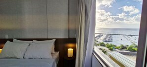 A hotel room with a bed, a nightstand with a lamp, and a view of a marina with boats. - CODDJ0100 - Cond. Nossa Sra de Lourdes (Salvador)