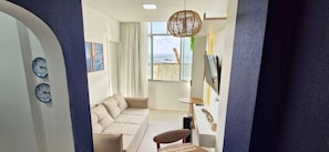 A modern living room with a sofa, a small table, a TV, and a view of the sea. - CODDJ0100 - Cond. Nossa Sra de Lourdes (Salvador)