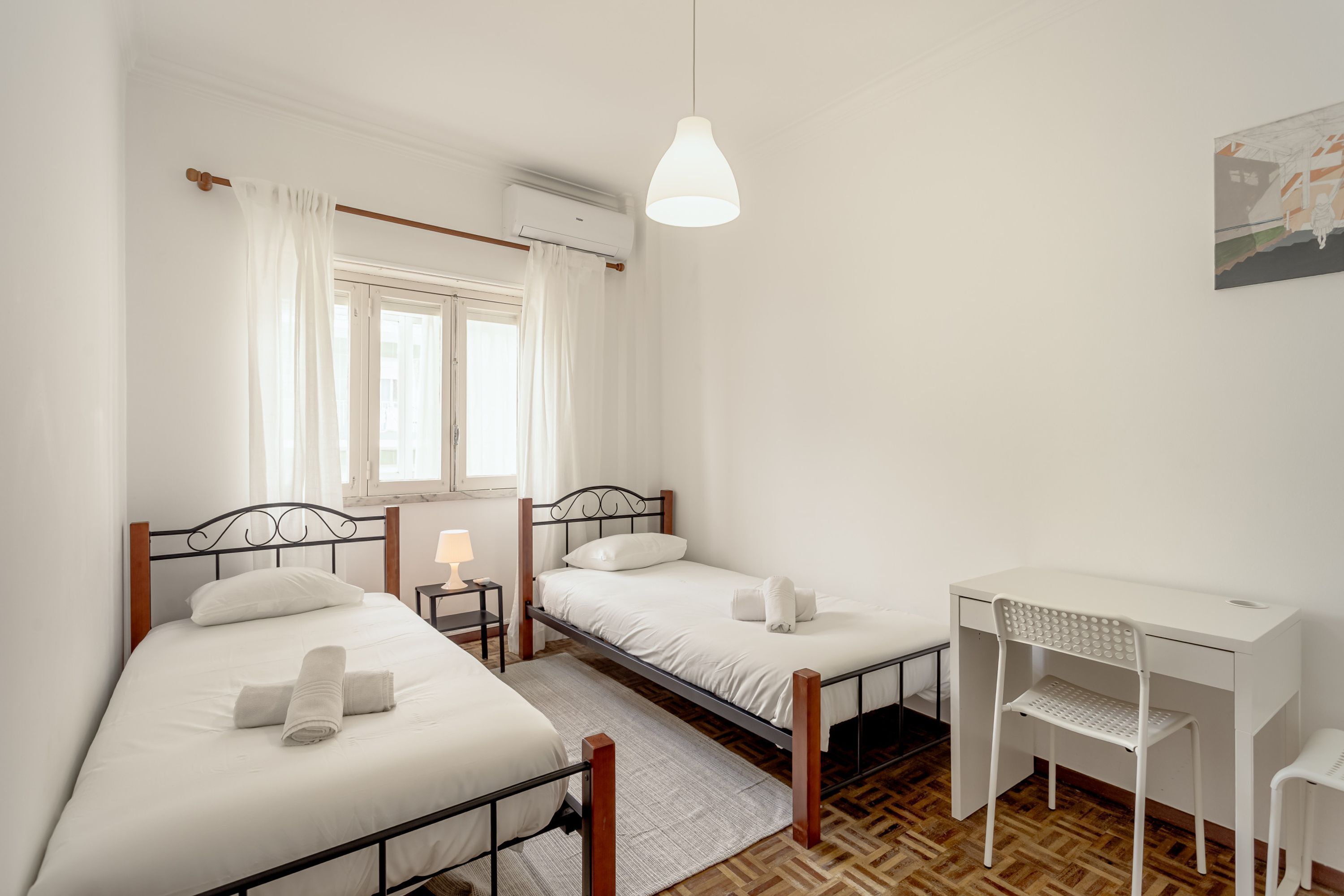 A bedroom with two beds, a desk, a chair, a lamp, and a painting on the wall.