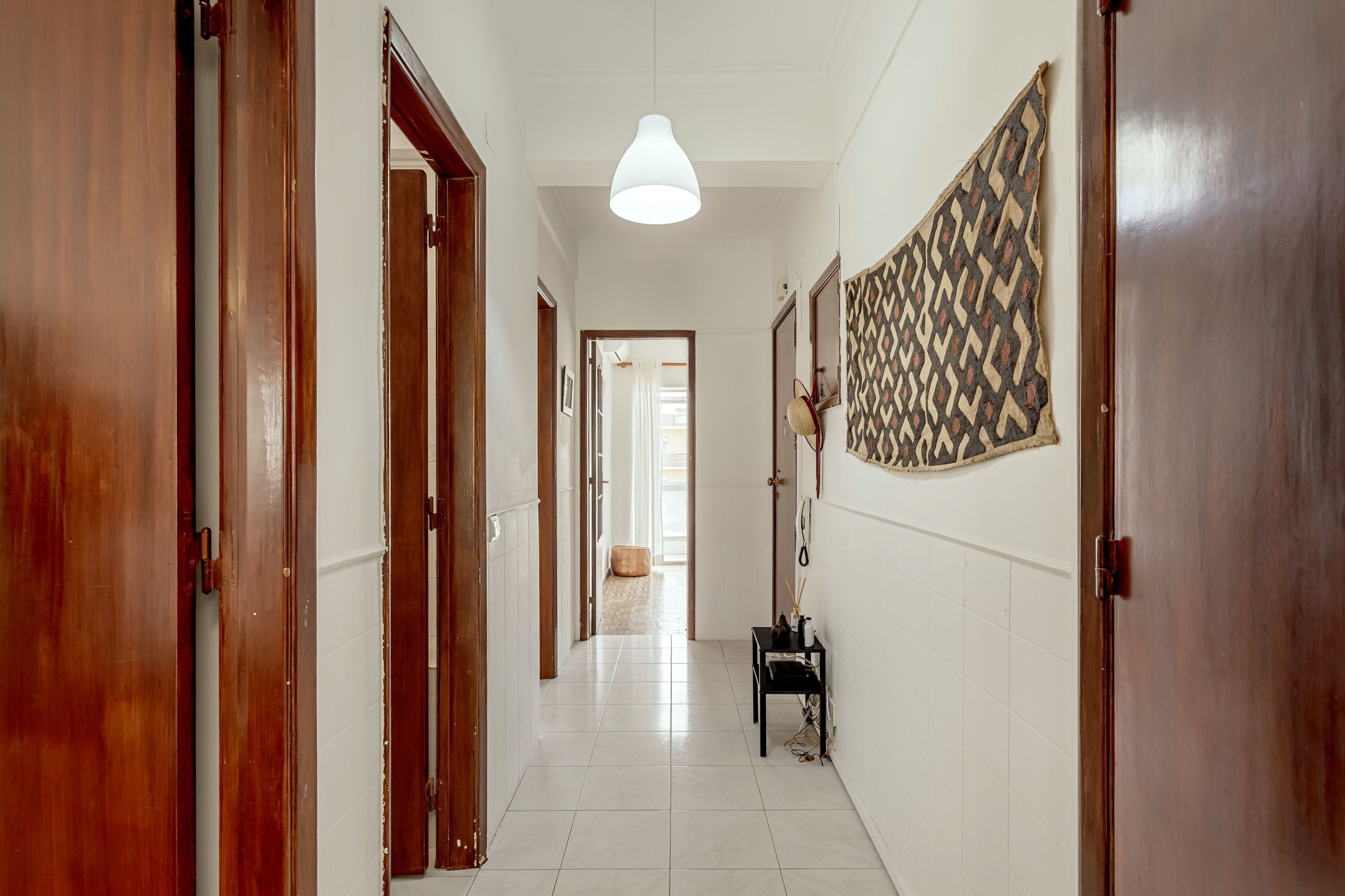 Un couloir avec des portes en bois, un luminaire suspendu, une décoration murale à motifs et une petite table sur laquelle est posé un vase.