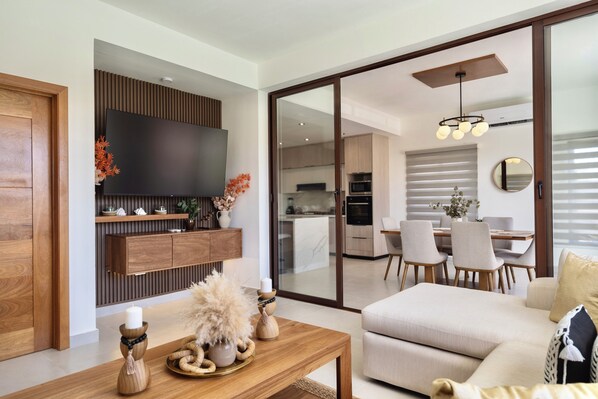 A modern living room with a flat-screen TV, wooden furniture, a dining area with chairs, and a kitchen in the background.