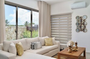 Un salón moderno con un sofá color beige, una mesa de centro de madera y vistas a la vegetación exterior.