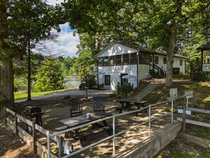 A picnic area with benches and a grill, surrounded by trees and a lake view.