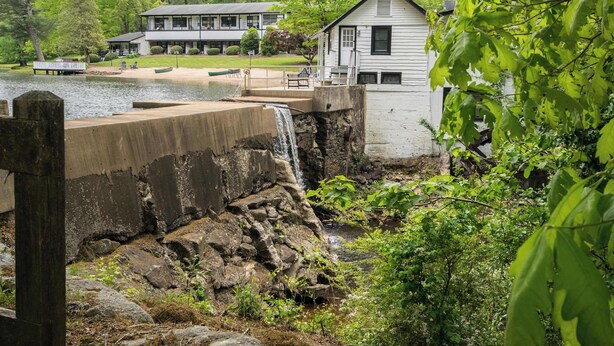 Uma casa à beira de um lago com uma cascata, rodeada por vegetação.