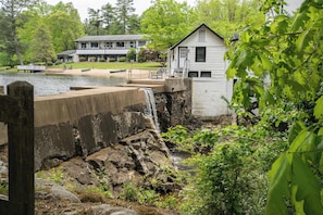 A house by a lake with a waterfall, surrounded by greenery.