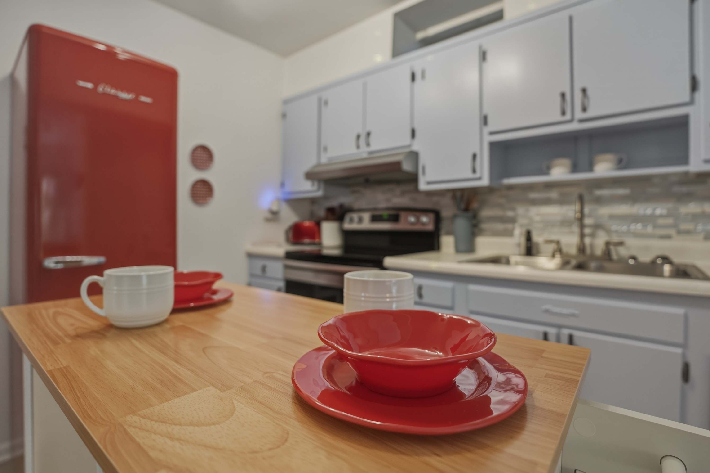 Une cuisine avec un réfrigérateur rouge, des armoires blanches et un plan de travail en bois, sur lequel se trouvent un saladier rouge et une tasse blanche.