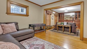 Ein Wohnzimmer mit einer braunen Ledersofa, einem Kicker, einem gemusterten Teppich, Holzböden, einem Fenster mit Holzrahmen und einer durch einen offenen Durchgang sichtbaren Küchenzeile.