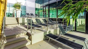 A modern building with a glass facade, outdoor seating area with sun loungers, and a staircase leading to an upper level.