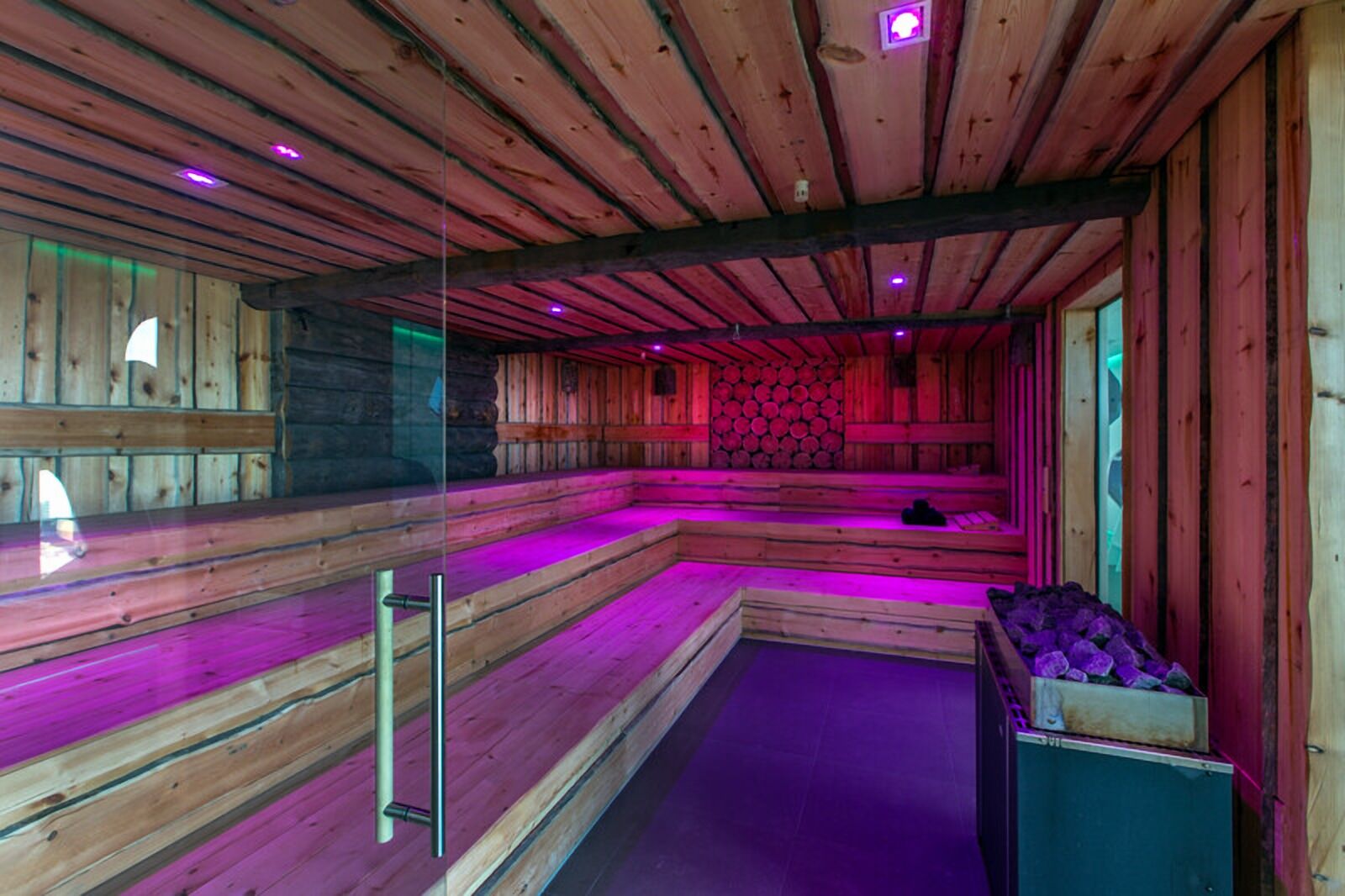 Una sauna con bancos de madera, pared de troncos, mampara de vidrio e iluminación morada.