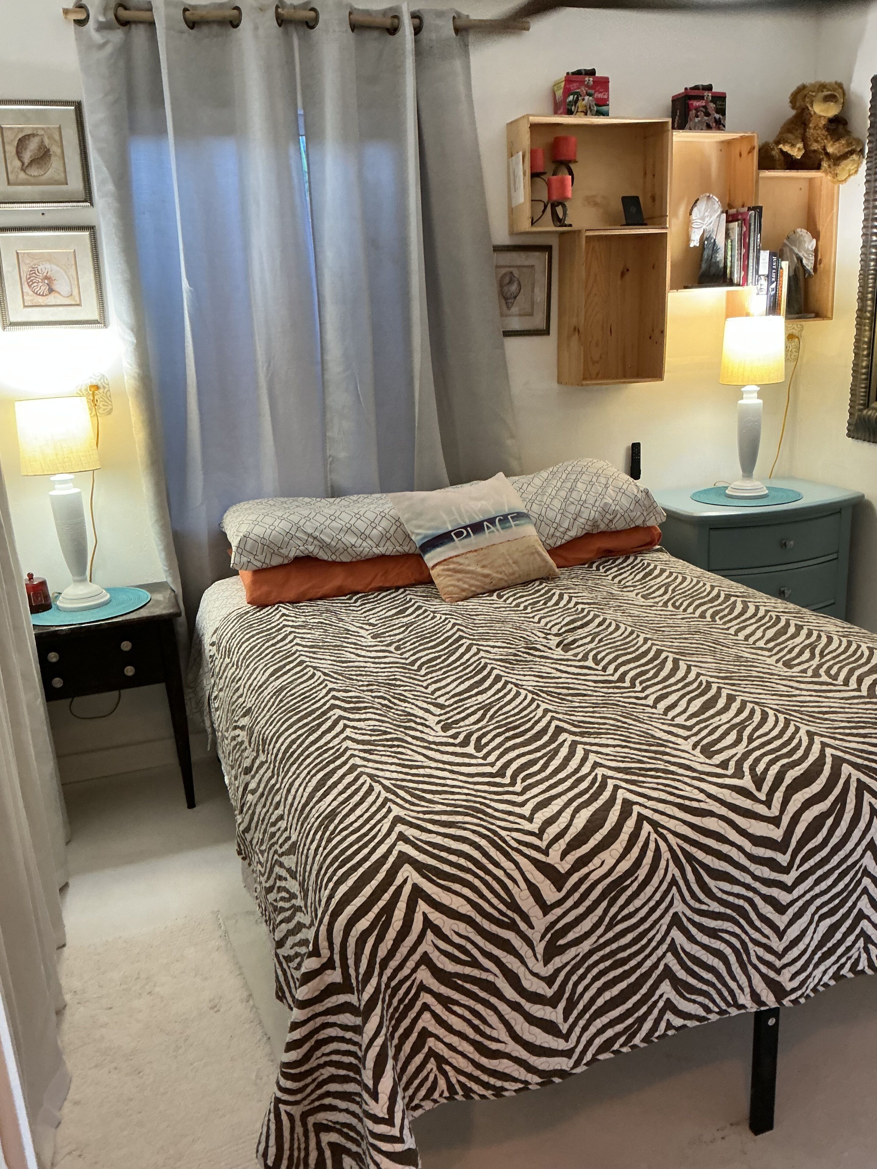 Een slaapkamer met een bedovertrek met zebraprint, een nachtkastje met een lamp, een plank met boeken en een teddybeer, en een gordijn.